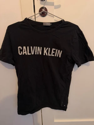 Svart Calvin Klein t-shirt - Svart t-shirt från Calvin Klein Jeans med vit logga tryckt över bröstet. Klassisk rund halsringning och korta ärmar. Tillverkad i mjuk bomull som är skön att bära. Perfekt för en stilren och enkel look.