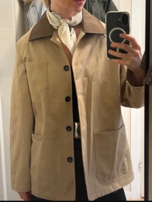 Beige jacka  - Stilren beige jacka med mörkbrun kontrastkrage och svarta knappar. Jackan har raka linjer, tre stora fickor framtill och är tillverkad i ett kraftigt bomullstyg. Perfekt för dig som gillar minimalistisk och tidlös design.