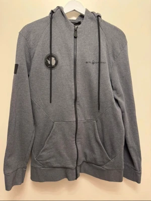 Grå zip-hoodie Sail Racing - Snygg grå hoodie från Sail Racing med dragkedja, huva med dragsko och två fickor framtill. Märkets logga finns på bröstet och ärmen samt en detalj på huvan. Mjuk bomullsblandning som passar perfekt till en avslappnad stil.