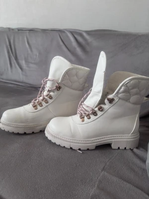 Vita quiltade boots med rosa detaljer - Snygga vita boots med quiltad vaddering runt ankeln och rosa skosnören. Grov sula och metallfärgade öljetter ger en cool look. Perfekta för dig som vill sticka ut med dina skor. Materialet ser ut att vara syntet och textil.