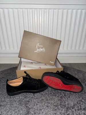 Svarta Christian Louboutin sneakers - Snygga svarta sneakers från Christian Louboutin med klassisk röd sula och coola nitar på tån. Skorna har snörning och är gjorda i mocka och läder. Perfekta för dig som vill sticka ut med exklusiv stil och detaljer som syns. Allt är bra med dom ba sulan kan man ba ta bort lite annars helt ny me dbox oxh allt med.