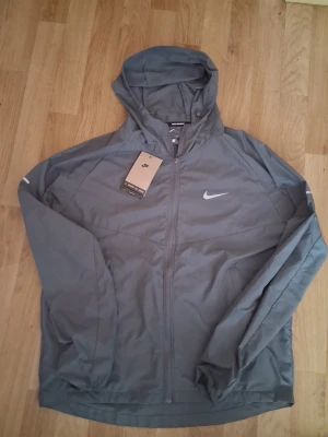 Grå vindjacka med huva från Nike - Snygg grå vindjacka från Nike med huva och dragkedja framtill. Jackan har en minimalistisk design med Nike-logga på bröstet och är tillverkad i lätt, vindtätt material. Perfekt för blåsiga dagar och sportiga outfits.