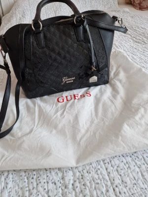 Svart handväska från Guess - Snygg svart handväska från Guess med präglat mönster och silverfärgade detaljer. Väskan har både handtag och avtagbar axelrem, samt dragkedja upptill. Guess-logga framtill och dekorativ tag. Perfekt för dig som vill ha en stilren och trendig accessoar.