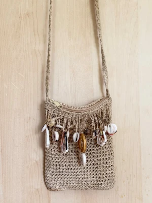 Beige virkad axelväska med snäckor - Trendig beige axelväska i virkat material med dekorativa snäckor och pärlor längs dragkedjan. Väskan har en smal, tvinnad axelrem och en fyrkantig form. Perfekt accessoar för en bohemisk look.