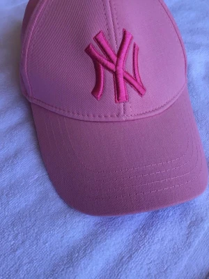 Rosa New York Yankees keps - Snygg rosa keps från New York Yankees med broderad NY-logga framtill i matchande rosa nyans. Klassisk böjd skärm och justerbar passform bak. Perfekt accessoar för en sportig och trendig look.
