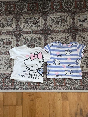 Hello Kitty t-shirts från H&M - Två söta Hello Kitty t-shirts från H&M. Ena t-shirten är vit med stor Hello Kitty-tryck och rosa rosett, den andra är blå- och vitrandig med flera Hello Kitty-motiv och rosa hjärtan. Båda har korta ärmar och är i mjuk bomull.