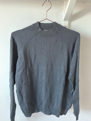 Grå stickad tröja från Zara - En stilren grå stickad tröja från Zara med högre ribbad krage,Tröjan har en enkel design utan mönster och är tillverkad i ett mjukt, tunt stickat material. Perfekt för lager-på-lager och passar till många olika outfits.
