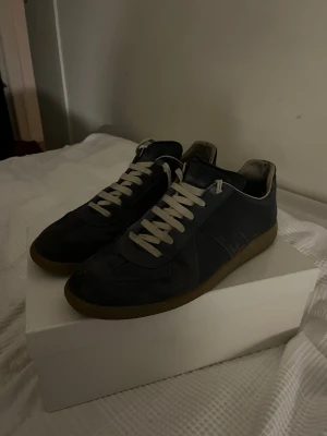 Mörkblå Maison Margiela Gats - Stilrena mörkblå Gats från Maison Margiela med klassisk låg siluett, vita snören och beige gummisula. Skorna har en minimalistisk design med diskreta paneler och är tillverkade i mocka och läder för en lyxig känsla. Köpta för cirka 1,5 år sen här på plick. Använda men fortfarande väldigt bra skick. Box, dustbag och kvitto medföljer. Storlek 45. Hör av dig vid minsta fundering!