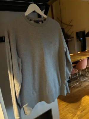 Grå sweatshirt från Gant - Klassisk grå sweatshirt från Gant med rund halsringning och diskret logga på bröstet. Tröjan har långa ärmar och ribbade muddar vid ärmslut och nederkant. Perfekt basplagg för en avslappnad stil.