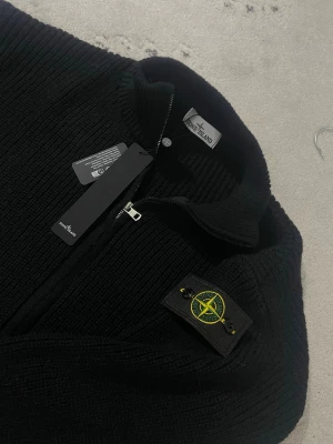  Stone Island - Säljer en svart stickad ziptröja från Stone Island med hög krage och ikonisk patch på ärmen. Tröjan har grovstickad struktur och dragkedja framtill. Perfekt för dig som gillar streetwear och vill ha något stilrent och bekvämt.