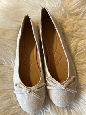 Beige ballerinaskor med rosett - Snygga ballerinaskor i beige tyg med rund tå och söt rosettdetalj framtill. Skorna har en mjuk innersula med diskret mönster och klassisk låg profil. Perfekta för en stilren och enkel look.