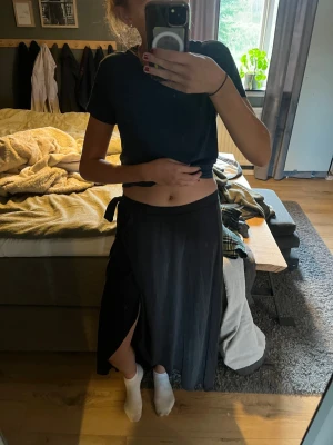 Svart omlottkjol med slits - Snygg svart långkjol i omlottmodell med slits på sidan. Kjolen har en enkel och stilren design som knyts i midjan och faller snyggt längs benen. Perfekt för dig som gillar en avslappnad men trendig look.