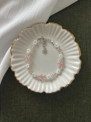 Sött armband med pärlor och rosa blommor - Armband med vita pärlor och rosa blommor i plast, trädd på metallkedja med justerbart lås. Blommorna är genomskinliga och ger en söt och romantisk känsla. Perfekt accessoar för att piffa upp din outfit.