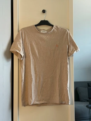 Beige t-shirt från Les Deux - Snygg beige t-shirt från Les Deux med liten broderad logga på bröstet. Klassisk rund halsringning och korta ärmar. Tillverkad i mjuk bomull som känns skön mot huden. Perfekt för en avslappnad och stilren look.