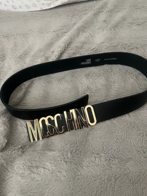 Svart skärp från Love Moschino - Stilrent svart skärp från Love Moschino med en stor, glansig guldfärgad logobuckle. Skärpet är tillverkat i skinn och har en klassisk, rak form som ger en cool och trendig look. Perfekt för att lyfta din outfit med en statement-accessoar.