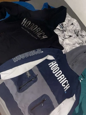 Två Hoodrich hoodies med tryck - Säljer två snygga hoodies från Hoodrich. En är helsvart i strl M med stort HOODRICH-tryck på bröstet, den andra är blå, grå och är i strl Xs men passar S vit med HOODRICH-tryck och två fickor med dragkedja framtill. Båda har huva och sportig streetwear-stil. Själv sö kan jag använda båda 2