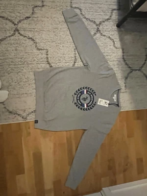 Grå sweatshirt från U.S. Polo Assn. - Snygg grå sweatshirt från U.S. Polo Assn. med rund halsringning och tryckt logga framtill i blått, vitt och rött. Tröjan har långa ärmar och ribbade muddar vid ärmslut och nederkant. Perfekt för en avslappnad och sportig stil. ÄKTA!