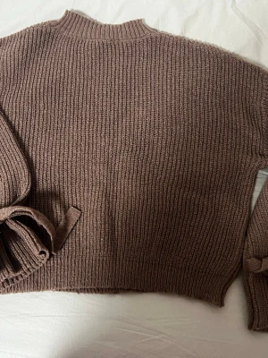 Brun ribbstickad tröja med knytband - Mysig brun ribbstickad tröja med långa ärmar och trendiga knytband vid ärmsluten. Tröjan har en något högre halsringning och är tillverkad i ett mjukt stickat material. Perfekt för lager-på-lager och chill dagar.
