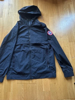 Svart vindjacka från Canada Goose - Svart vindjacka från Canada Goose med huva och dragkedja framtill. Jackan har två sidofickor med dragkedja och den klassiska runda Canada Goose-loggan på vänster ärm. Lätt och smidig modell i tunt material, perfekt för blåsiga dagar.