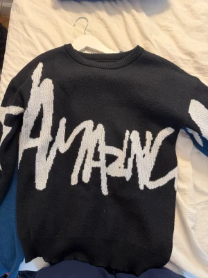 Svart stickad tröja med vit text - Cool svart stickad tröja med stor vit text i graffiti-stil som säger 'AMAZING' över hela framsidan. Tröjan har rund halsringning och långa ärmar. Perfekt för dig som gillar streetwear och vill sticka ut med en unik look.