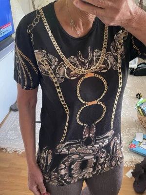 Svart t-shirt med kedjemönster - Cool svart t-shirt med tryck av guldfärgade kedjor och ormliknande detaljer. T-shirten har rund halsringning och korta ärmar. Perfekt för dig som vill sticka ut med ett unikt och trendigt mönster.