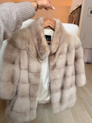 Beige pälsjacka med bred krage - Lyxig beige pälsjacka från EF med bred krage och markerade horisontella sömmar. Jackan har en mjuk, fluffig yta och är helfodrad i ljust tyg. Perfekt för att hålla värmen under kalla vinterdagar.