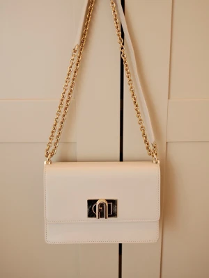 Beige handväska med guldkedja - Stilren beige handväska i slätt läder med guldfärgad kedjerem och elegant metallspänne framtill. Väskan har en klassisk rektangulär form och lock med magnetlås. Perfekt för dig som gillar minimalistisk och tidlös design.