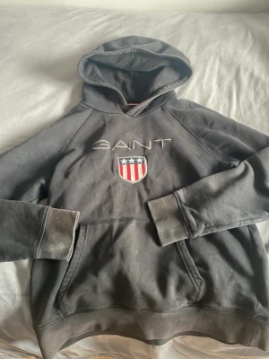 Svart hoodie från GANT med logga - Svart hoodie från GANT med broderad logga och amerikanskt inspirerat sköldmärke på bröstet. Klassisk känguruficka framtill och huva med dragsko. Mjuk bomullskvalitet och ribbade muddar vid ärmslut och nederkant. Perfekt för en avslappnad och sportig stil.