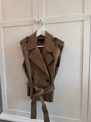 Beige trenchväst från ZARA - Stilren beige trenchväst från ZARA med breda slag, knappar och knytskärp i midjan. Västen har axelklaffar och är ärmlös, perfekt för lager-på-lager-look. Materialet är mjukt och följsamt, vilket ger en snygg siluett. Helt ny med prislapp kvar 