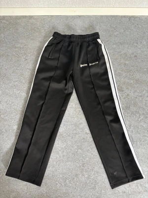Palm Angels svarta mjukisbyxor - Svarta mjukisbyxor från Palm Angels med vita sidoränder och logga på vänster ben. Byxorna har resår i midjan och raka ben. Perfekta för en avslappnad och trendig stil.