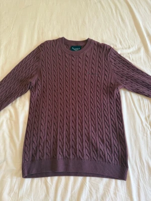 Bondelid Cable Knit - Säljer en plum färgad cable knit tröja från Bondelid med rund halsringning och långa ärmar. Tröjan har ett klassiskt kabelmönster och ribbade muddar vid ärmslut och nederkant. Perfekt för dig som gillar en stilren och tidlös look.