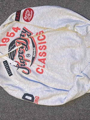Superdry grå hoodie med tryck - Snygg grå hoodie från Superdry med stora röda och svarta tryck på bröstet och ärmen. Hoodien har huva med dragsko, känguruficka och coola broderade detaljer som 'Racing' och 'Classics 1954'. Perfekt för en avslappnad streetstyle-look.