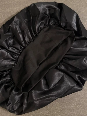 Svart satin hårbonnet - Säljer en svart hårbonnet i glansigt satinmaterial. Perfekt för att skydda håret under natten och minska friss. Bonneten har en elastisk kant som gör att den sitter bekvämt på huvudet. Enkel och stilren design som passar alla hårtyper.