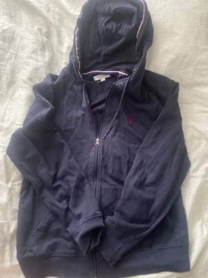 Marinblå hoodie från US Polo Assn - Mörkblå hoodie med dragkedja från US Polo Assn. Klassisk design med röd logga broderad på bröstet och vit-röd randig detalj i huvan. Mjuk bomullskänsla och fickor framtill. Perfekt för en chill och sportig stil.