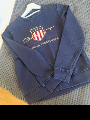 Blå sweatshirt från Gant - Mörkblå sweatshirt från Gant med broderad logga och texten 'American Sportswear' framtill. Tröjan har rund halsringning, långa ärmar och ribbade muddar vid ärmslut och nederkant. Perfekt för en avslappnad och sportig look.