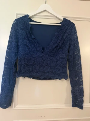 Blå spets topp med v-ringning - Snygg blå långärmad topp i spets med v-ringad hals och croppad passform. Toppens spetsmönster ger en romantisk vibe och den har vågig kant nedtill. Perfekt att styla med höga jeans eller kjol.