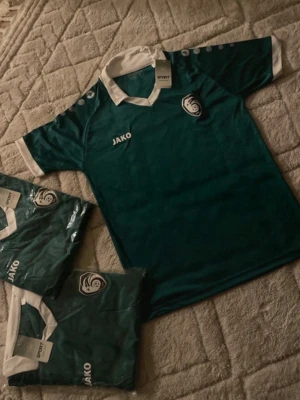 Grön fotbollströja från Jako - Snygg grön fotbollströja från Jako med vitt krage och vita detaljer vid ärmslut. Tröjan har klubbmärke på bröstet och Jako-logga. Perfekt för träning eller match.