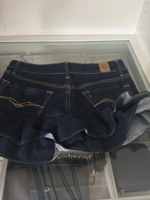Mörkblå jeanskjol med volang(waist=25) - Säljer en mörkblå jeanskjol från Levi's med volang nedtill och klassiska fickor både fram och bak. Kjolen har kontrastsömmar och en snygg, kort passform som ger en cool och ungdomlig look. Perfekt för dig som gillar denim och vill sticka ut.