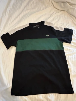Svart och grön t-shirt från Lacoste - Snygg svart t-shirt från Lacoste med en bred grön rand över bröstet och klassisk krokodillogga. T-shirten har rund halsringning och korta ärmar. Materialet är mjuk bomull och modellen är normal i passformen.