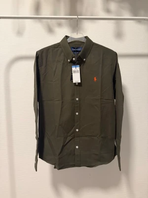 Olivgrön skjorta från Ralph Lauren - Klassisk olivgrön skjorta från Ralph Lauren med orange broderad logga på bröstet. Skjortan har knappar framtill, button-down krage och långa ärmar. Tillverkad i mjuk bomull för en bekväm känsla och stilren look.
