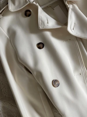 Klassisk vit trenchcoat med knappar - Stilren vit trenchcoat med dubbelknäppning och stora bruna knappar. Jackan har bred krage och bälte i midjan för en snygg siluett. Perfekt för dig som gillar tidlös och clean stil. Materialet känns slätt och kvalitativt.
