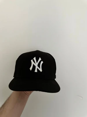 Svart New York Yankees keps - Snygg svart keps från New Era med klassisk New York Yankees-logga broderad i vitt framtill. Kepsen har rak skärm och justerbar passform. Perfekt accessoar för en sportig och urban stil.