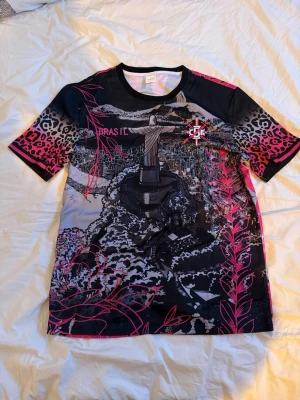Svart och rosa Brasilien t-shirt - Unik t-shirt med svart bas, rosa detaljer och leopardmönster på ärmarna. Stort tryck av Kristusstatyn i Rio och stadsmotiv både fram och bak. Kortärmad och tillverkad i ett lätt, syntetiskt material. Perfekt för dig som vill sticka ut med en cool streetstyle. Storleken är L, axellmått 48cm, längd 72cm.