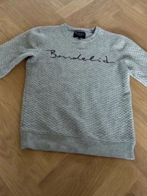 Grå sweatshirt från Bondelid - Snygg grå sweatshirt från Bondelid med våfflad struktur och svart broderad logga över bröstet. Tröjan har rund halsringning, långa ärmar och ribbade muddar vid ärmslut och nederkant. Perfekt för en avslappnad och stilren look.