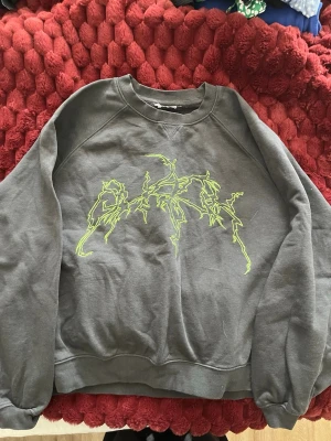 Svart sweatshirt med neontryck - Cool svart sweatshirt med ett stort, grafiskt tryck i neon-grönt framtill. Tröjan har rund halsringning, långa ärmar och ribbade muddar vid ärmslut och nederkant. Perfekt för dig som gillar streetwear