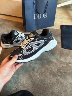 Svarta Dior sneakers med vita detaljer - Snygga svarta Dior sneakers med vita och grå detaljer samt vita skosnören. Skorna har en chunky sula och mesh- och läderpaneler för en modern look. Dior-logga syns på sidan och tungan. Perfekta för dig som vill sticka ut med exklusiva sneakers.