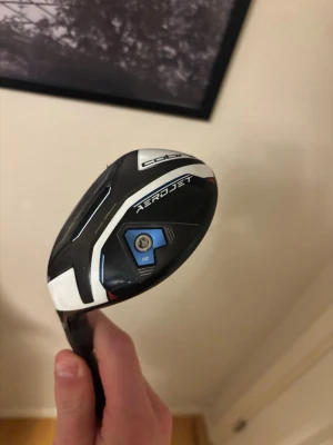 Cobra Aerojet hybrid 4 - Säljer en Cobra Aerojet hybrid golfklubba i svart, vitt och blått. Klubban har en modern design med tydliga linjer och justerbar vikt. Perfekt för dig som vill ha extra kontroll och längd på golfbanan. Den är i princip nyskick och nypris på denna är 2600kr  obs vänsterklubba