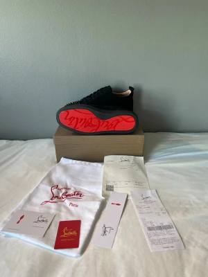 Christian Louboutin svarta sneakers - Säljer ett par svarta sneakers från Christian Louboutin med klassisk röd sula och svarta nitar över tån. Skorna är låga och har en lyxig känsla med mockaliknande material och svarta skosnören. Perfekta för dig som vill sticka ut med exklusiv design.