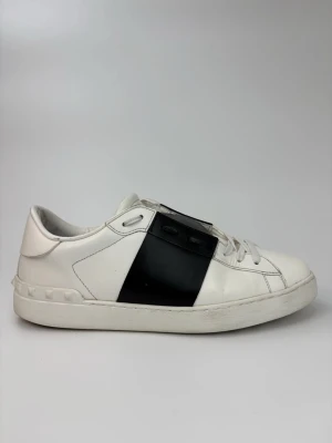 Valentino open skor - Snygga vita sneakers från Valentino med en bred svart läderdetalj över mitten. Skorna har klassisk låg profil, vita snören och diskreta nitar på hälen. Tillverkade i läder för en lyxig känsla och stilren look.