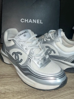 Chanel runners - Snygga sneakers från Chanel i vitt och silver med ikoniska CC-loggan på sidan. Skorna har en chunky sula, mesh- och läderdetaljer samt vita skosnören. Perfekta för dig som vill sticka ut med exklusiv stil och streetkänsla. Skorna beställdes i storlek 44 men sitter som storlek 40-41 så rekommenderar att du som köper har 40-41 i skostorlek